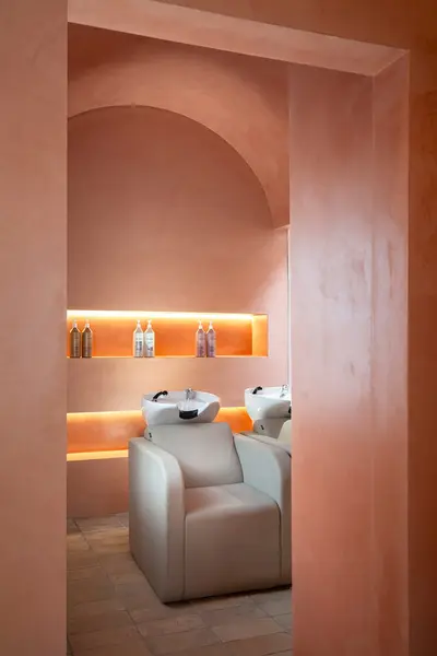 Backwash alcove seen through an arched opening, two basins and beige chairs below a backlit niche of product
