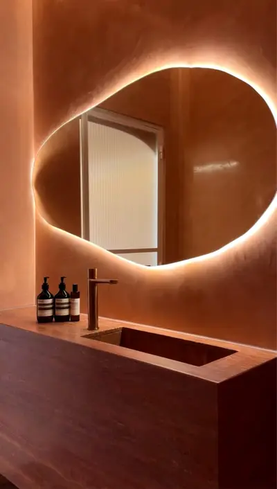Powder room with a pebble-shaped backlit mirror on terracotta plaster above a dark timber vanity with an integrated basin and brass tap