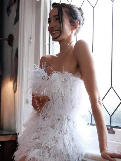 Woman in a white ostrich-feather mini dress laughing by a leadlight window, dark hair in a soft up-do with face-framing strands