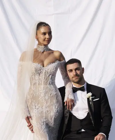Bride in a lace gown, choker and long veil beside the groom in a black tuxedo, against a white backdrop