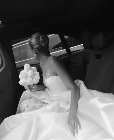Black-and-white photograph of a bride seated in a car, hair in a smooth low bun, holding white hydrangeas