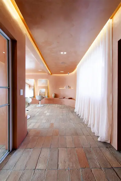 Corridor of terracotta pavers running past a full-height sheer curtain, under a coved ceiling washed with warm light, towards the styling chairs
