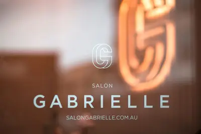 The shopfront window, etched with the Salon Gabrielle monogram and web address, with the neon monogram lit inside