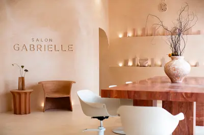 Lounge area in clay plaster, with Salon Gabrielle lettering set into the wall, a tan leather armchair and a red marble table with white swivel chairs