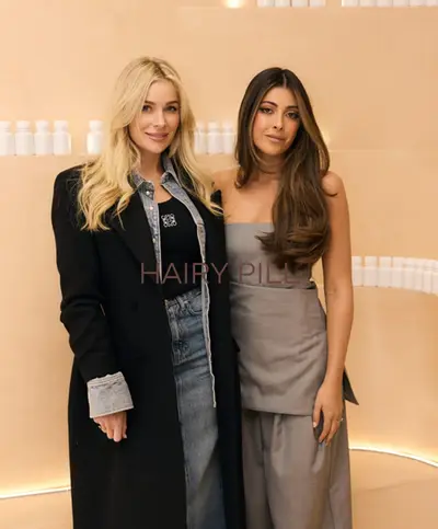 Two guests at a product launch in the salon, one in a black blazer over denim and one in a grey strapless dress, standing in front of a shelf of white bottles