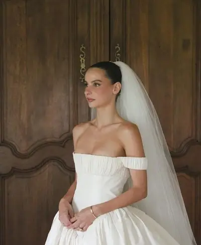 Bride in an off-the-shoulder ivory gown and long veil, hair in a sleek low bun, in front of carved wooden doors