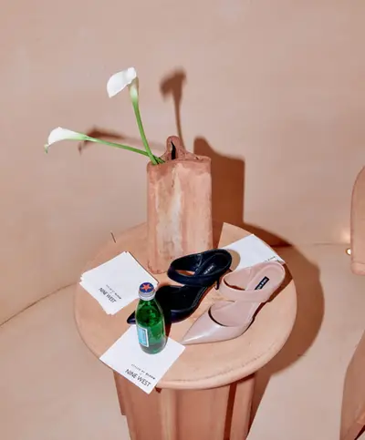 Two pairs of shoes and a bottle arranged on a round stone table beneath a vase of white calla lilies