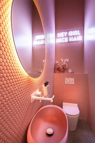 Powder room lined in pink penny-round tile, with a round backlit mirror, a conical pink basin and a neon sign reading Hey Girl Nice Hair