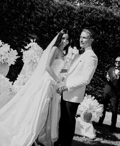 Black-and-white photograph of a bride in a strapless gown and long veil holding hands with the groom among white flowers