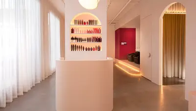 Curved cream reception desk beneath an arched niche of backlit retail shelves, with sheer floor-length curtains and a polished concrete floor