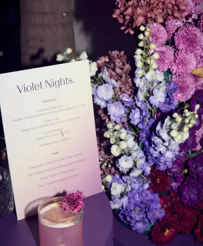A printed cocktail menu headed Violet Nights propped among dense purple and lilac flowers